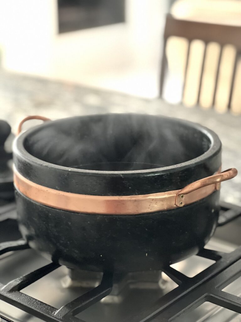 Handcrafted Brazilian soapstone cooking pot with copper band handles simmering on a gas stovetop, releasing steam in a modern kitchen setting.