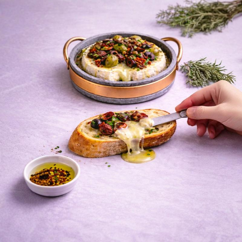 Soapstone brie pan with copper handles serving baked brie cheese with olives and herbs, natural stone cookware for oven baking